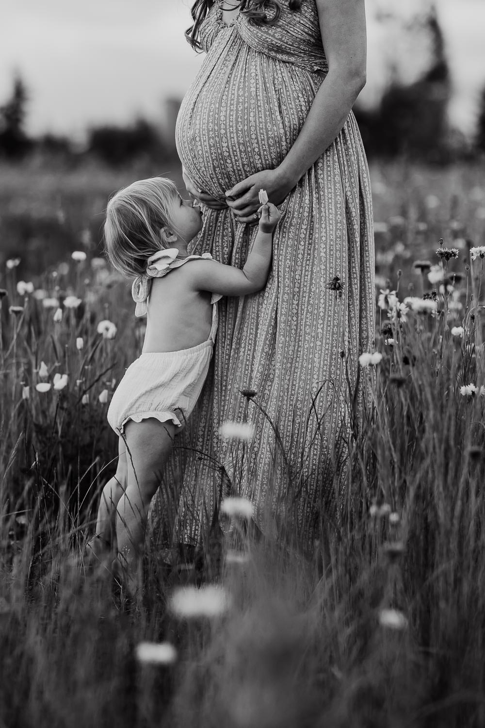 black and white image of toddler girl trying to kiss mommy's pregnant belly but she isn't tall enough