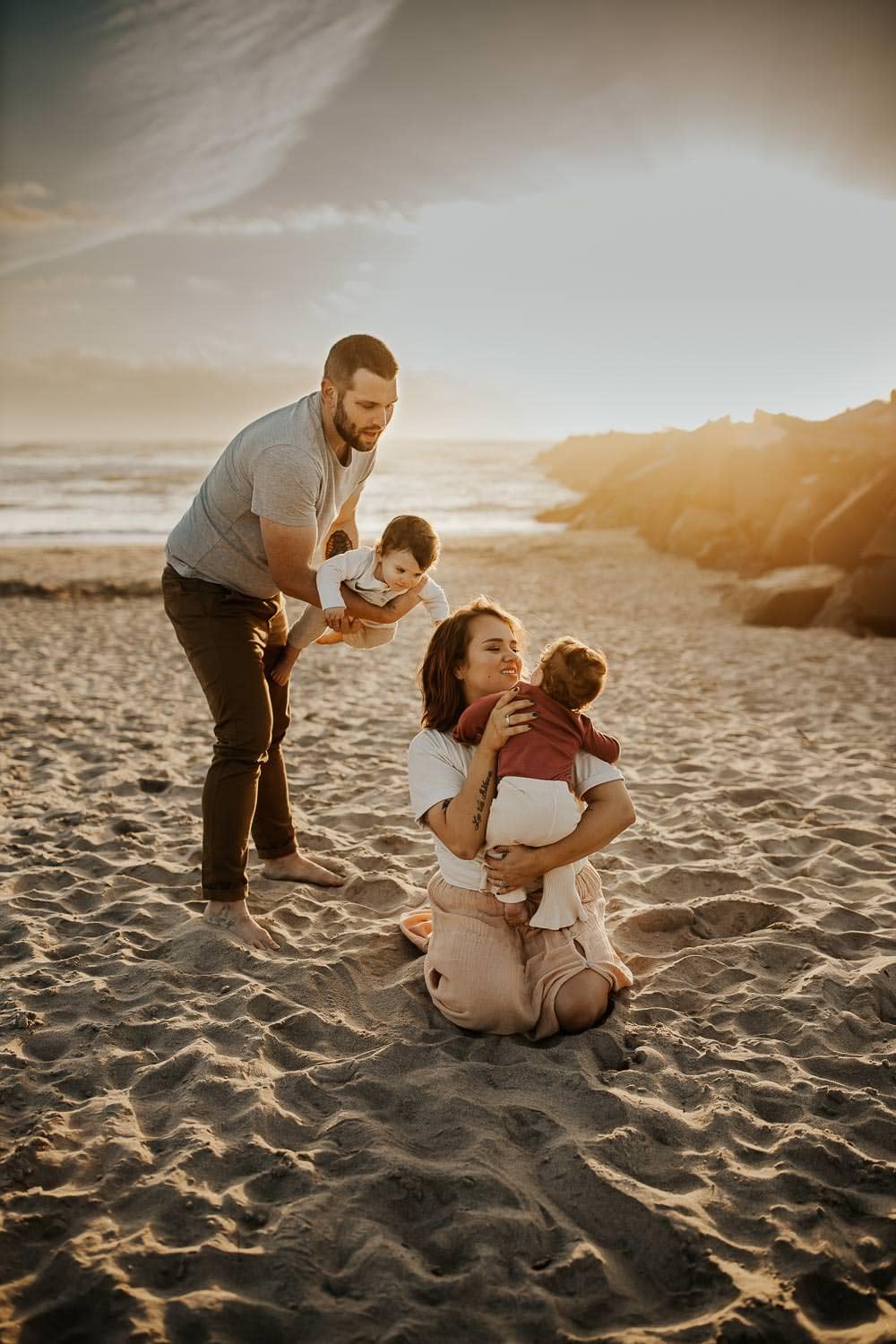 family photographer on the oregon coast near Manzanita - Family of four, mom sitting on sand holding baby girl, dad standing playing airplane with baby boy.