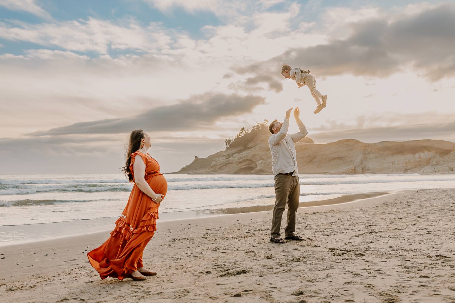 Dad throwing toddler in the air while pregnant mom watches at the oregon coast