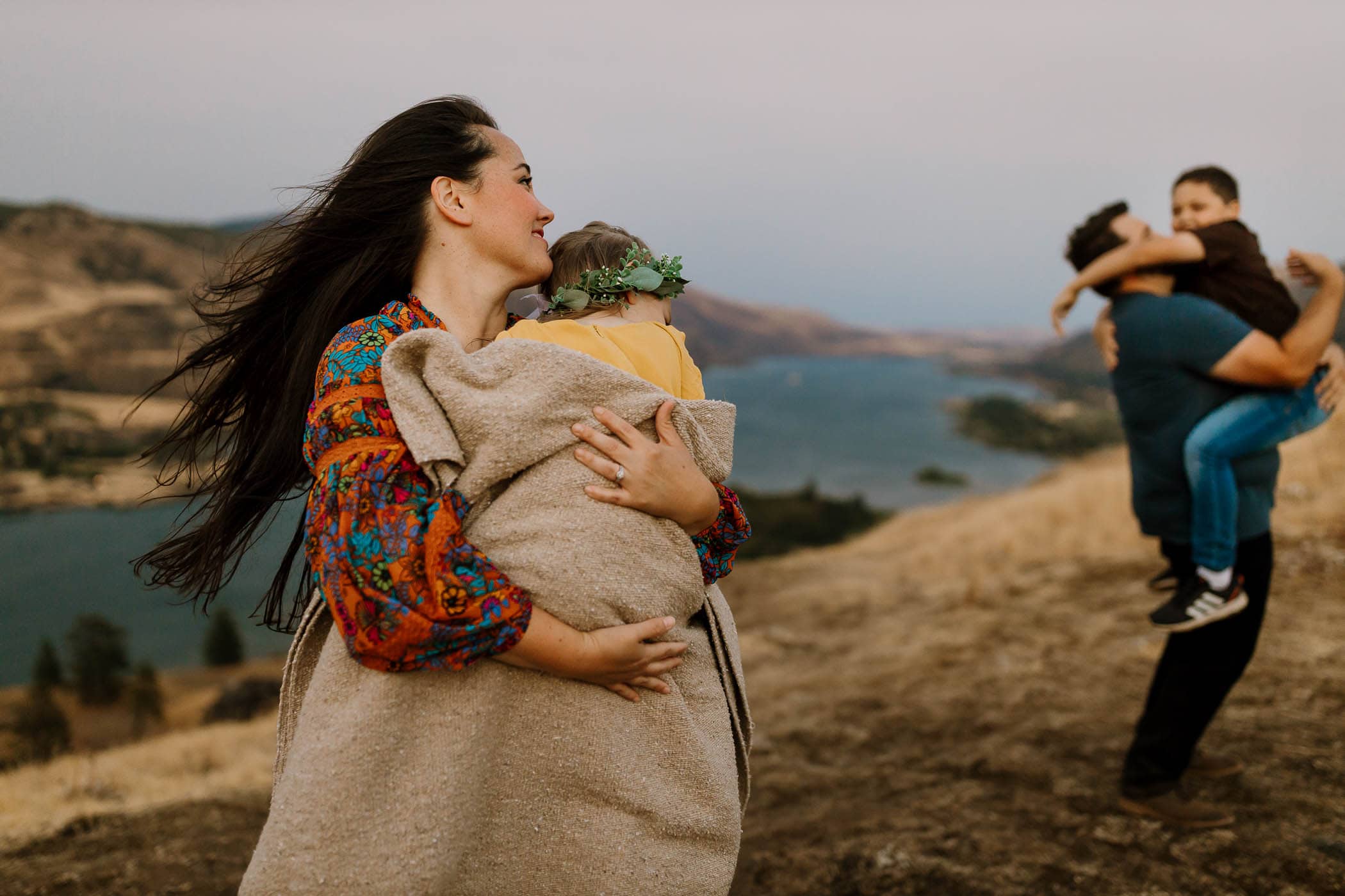 Mom holding daughter wrapped in blanket and looking back at dad and son playing - hood river family photography