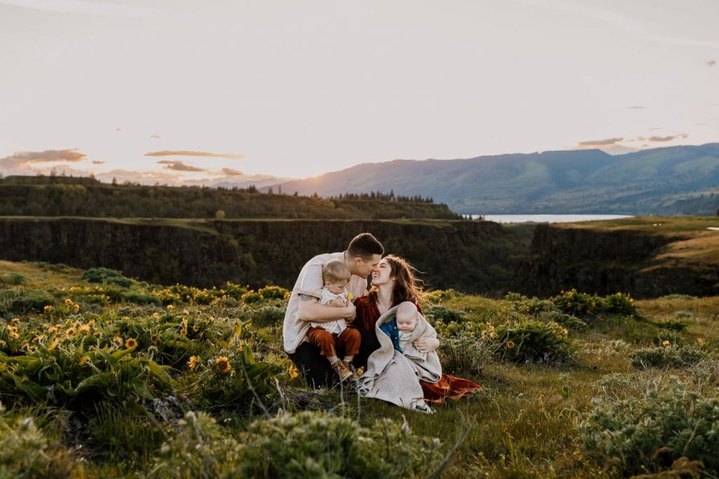 Mom and dad almost kiss while holding kids on their laps in the wildflowers near Portland Oregon