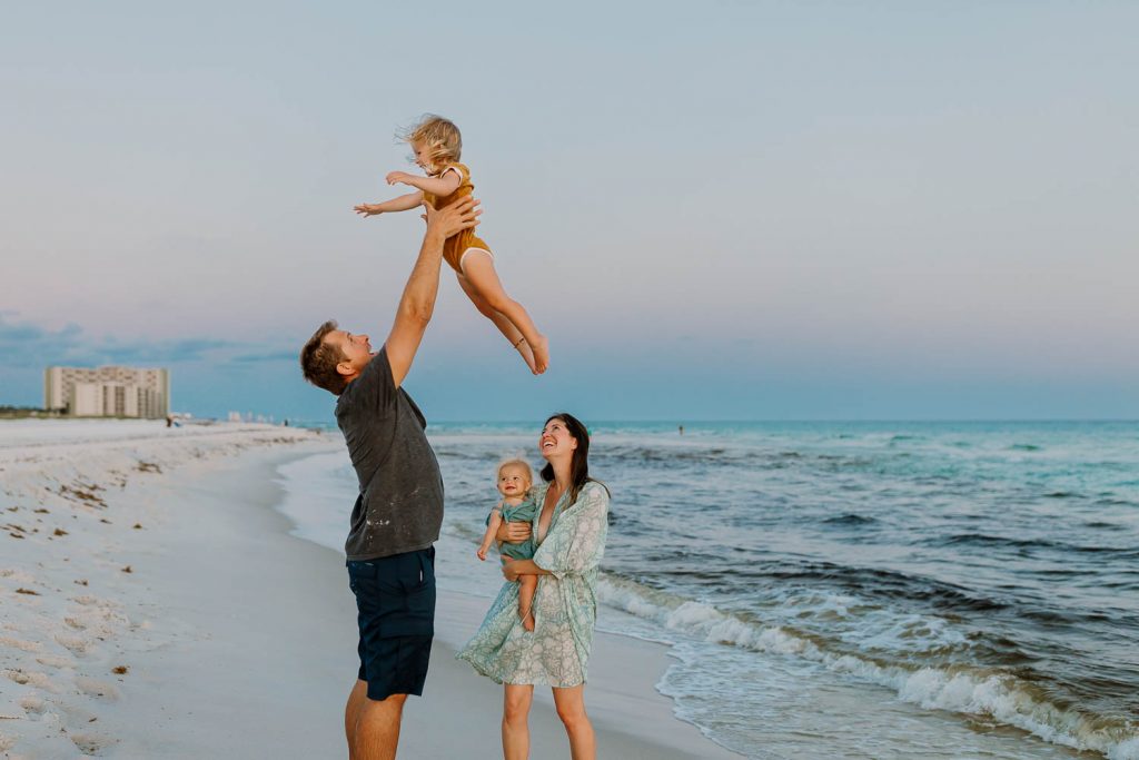 Dad throwing daughter in the air at the beach in Florida while mom and baby watch during travel family photos