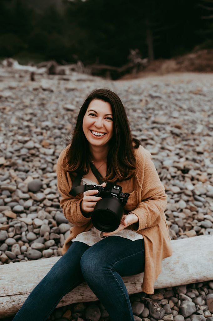 A family photographer sitting on a log and smiling while holding a Canon EOS R mirrorless camera