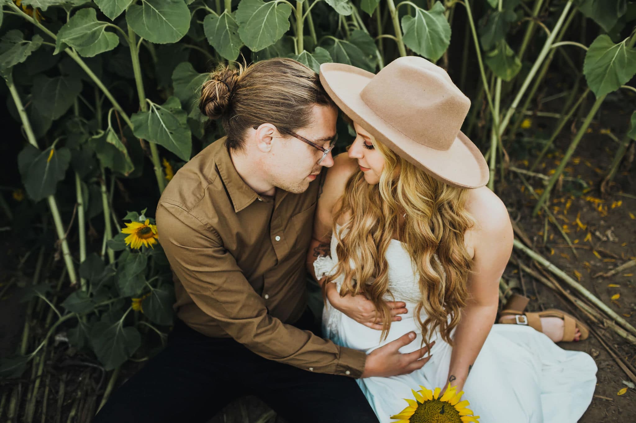 Pregnant mom wearing white dress and straw hat while husband hugs her