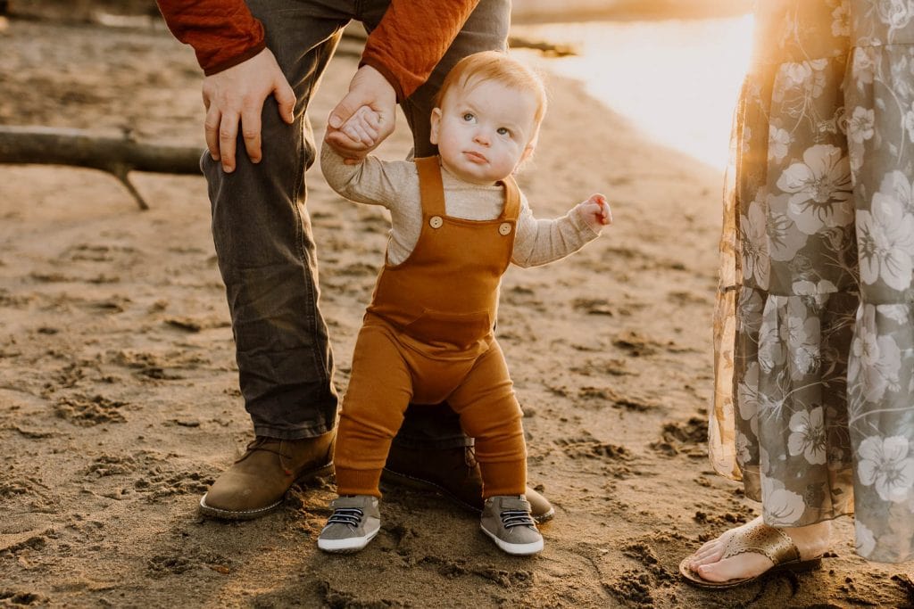 little baby boy standing holding dads hand, wearing brown overalls
