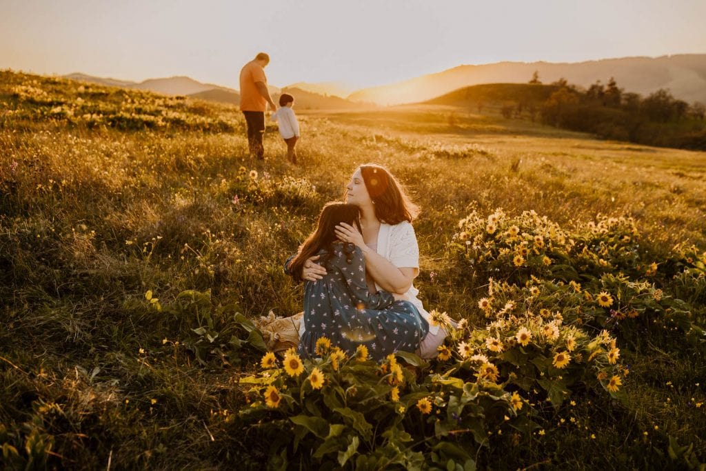 mother cuddling daughter in wildflowers while dad and son play behind them during sunset time