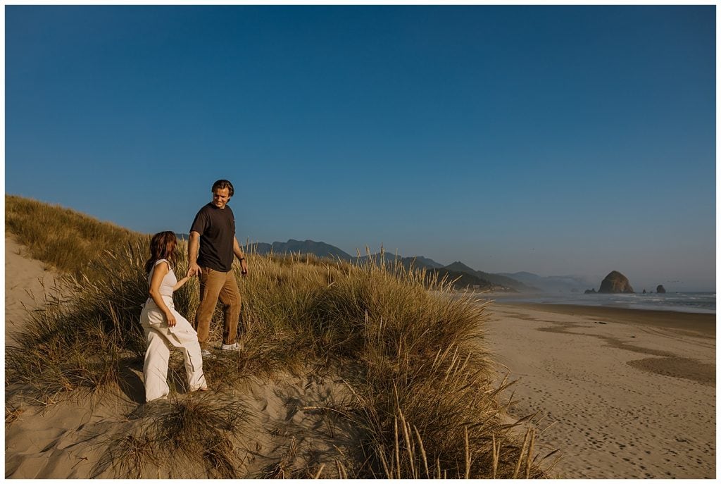 engaged couple walking up a sand dune in cannon beach oregon
