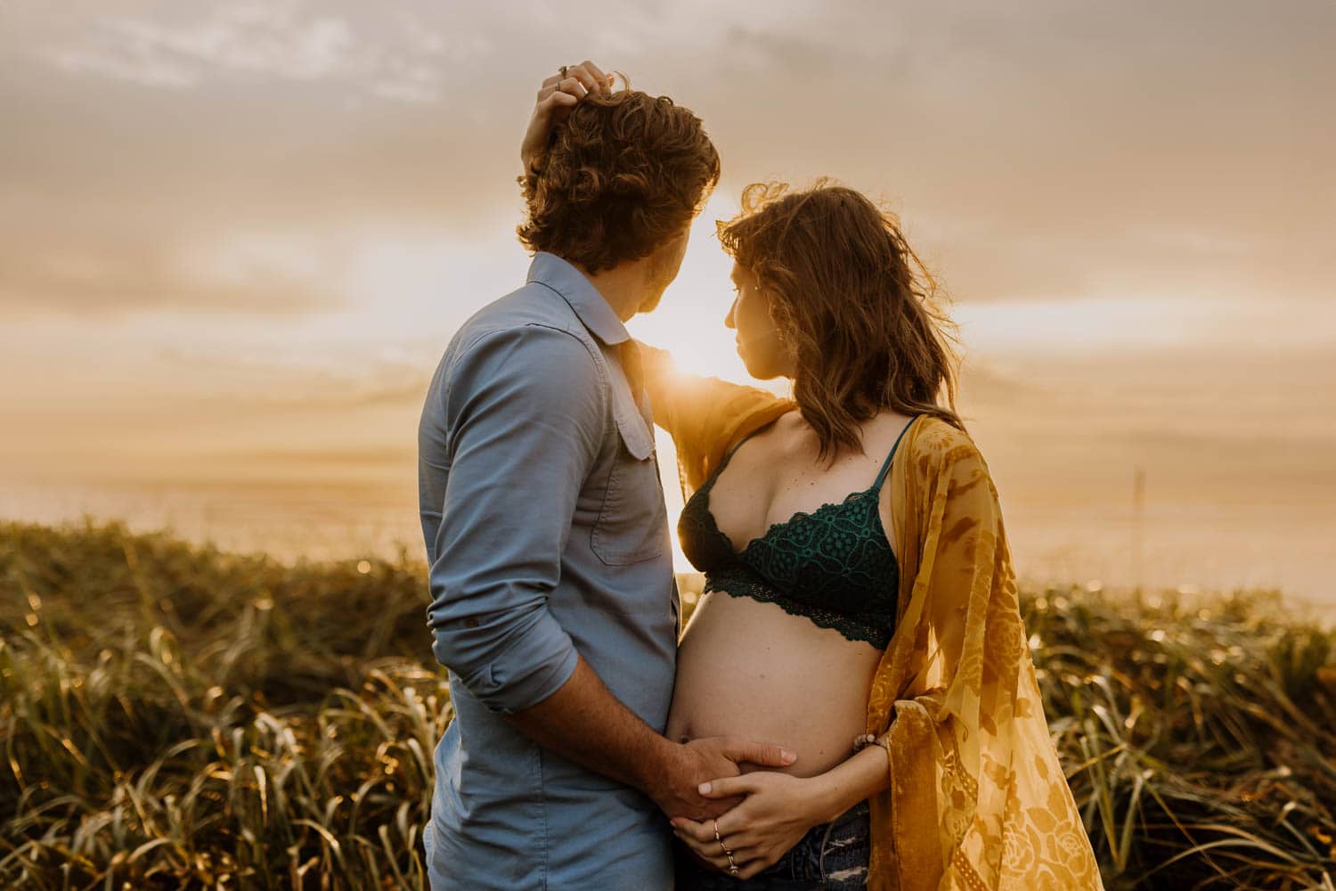 married couple holding each other close with bare pregnant belly and sunset