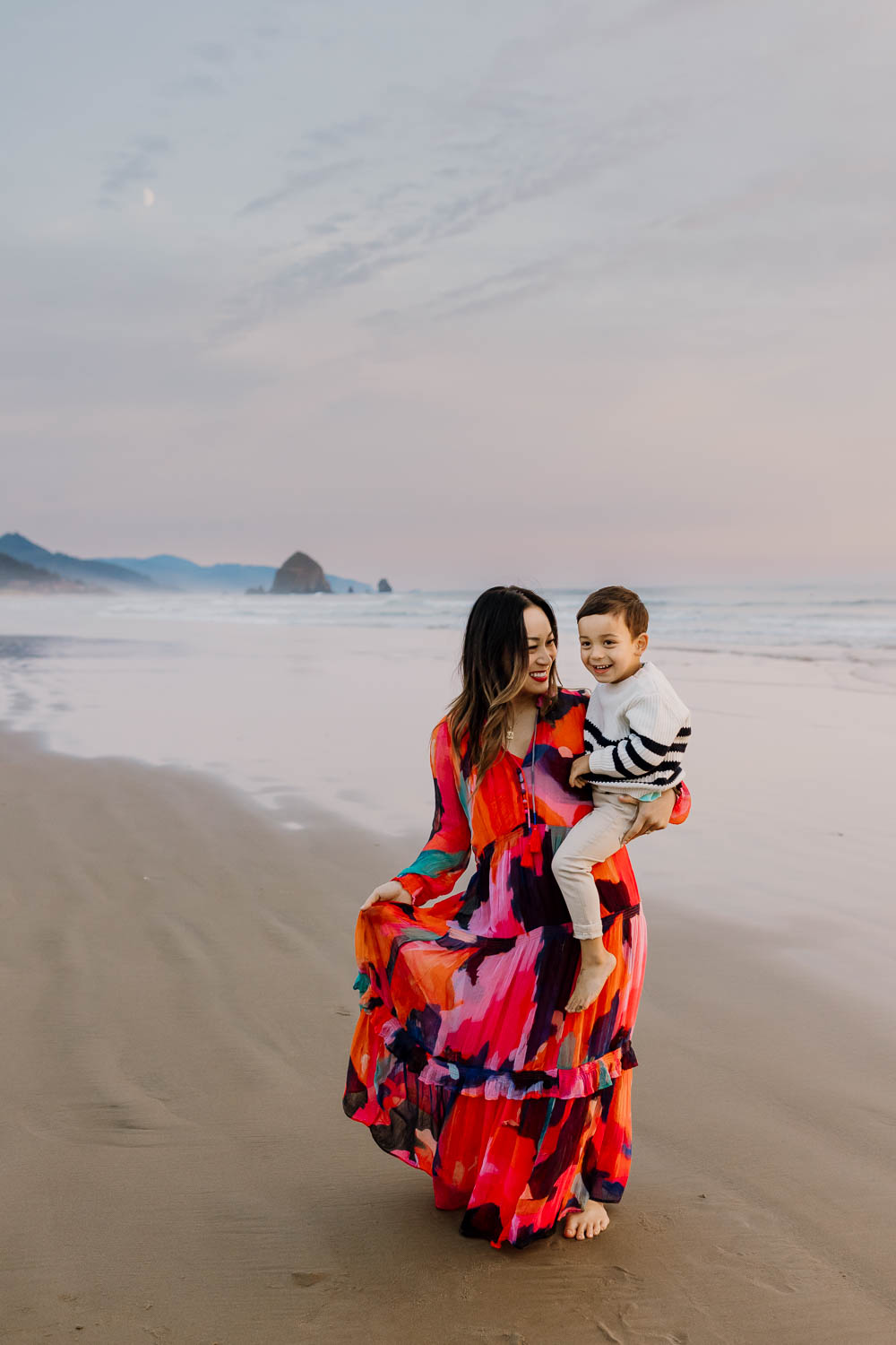 Mom in bright patterned dress holding son with Haystack Rock in cannon beach behind them